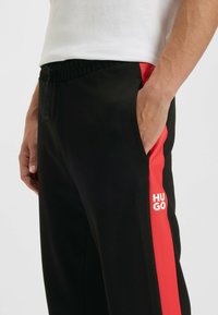 Sorte sportsbukser med rød sidestreg og hvid "HUGO" logo. Har elastisk talje og sidelommer. Glat stofstruktur.