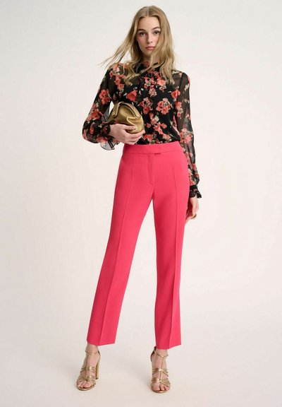 Pantalones rosa ajustados con cintura alta y costuras laterales, combinados con una blusa negra de flores transparente. Un bolso de mano dorado y tacones con tiras completan el look.