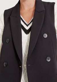 Czarna bezrękawnikowa marynarka z dwoma dużymi guzikami, klapami w kształcie litery V i gładką teksturą, nałożona na biały sweter z czarnym wzorem w kształcie litery V.