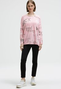 Junge Frau steht, trägt ein rosa Langarmshirt mit schwarzen Textdruck, schwarze Hose und weiße Sneakers vor einem schlichten weißen Hintergrund.