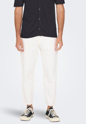 Pantalones de algodón blancos, diseño ajustado, de longitud corta, combinados con zapatillas de lona negras. Camisa oscura de botones de manga corta.