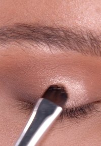 Oogschaduw aanbrengen met een platte kwast op een lichtbeige ooglid, met een zachte shimmer effect. De achtergrond toont de huidtextuur en de wenkbrauw.