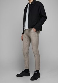 Svart jacka över en vit skjorta, ljusbeige slim-fit byxor och svarta låga sneakers, vilket visar en avslappnad och modern outfit.