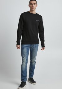 Black sweater with crew neck, textured fabric, featuring a small logo patch on the chest, paired with light blue jeans and black sneakers.