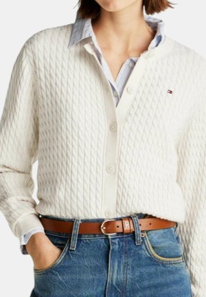 Kvinde iført hvid tekstureret cardigan over en blå skjorte med krave, stoppet ned i blå jeans med et brunt læderbælte.