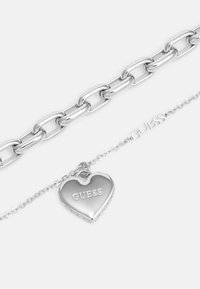 Bracelet en chaîne en argent avec des maillons allongés. Présente un pendentif en forme de cœur avec "GUESS" gravé, accompagné d'une délicate chaîne.