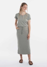 Grijze, lichte, mouwloze jurk met een trekkoord in de taille, zijzakken en spltjes. Draagt bij bruine sandalen. Gladde textuur.