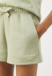 Calções de algodão numa tonalidade verde-limão com um cós elástico, bolsos laterais e detalhe em logótipo dourado na perna esquerda. Textura suave, design casual.