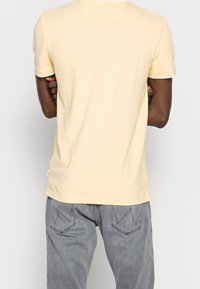 Kortärmad gul T-shirt i ett mjukt, slätt material; figursydd design med rundad halsringning och sidofickor för komfort. Grå byxor synliga.