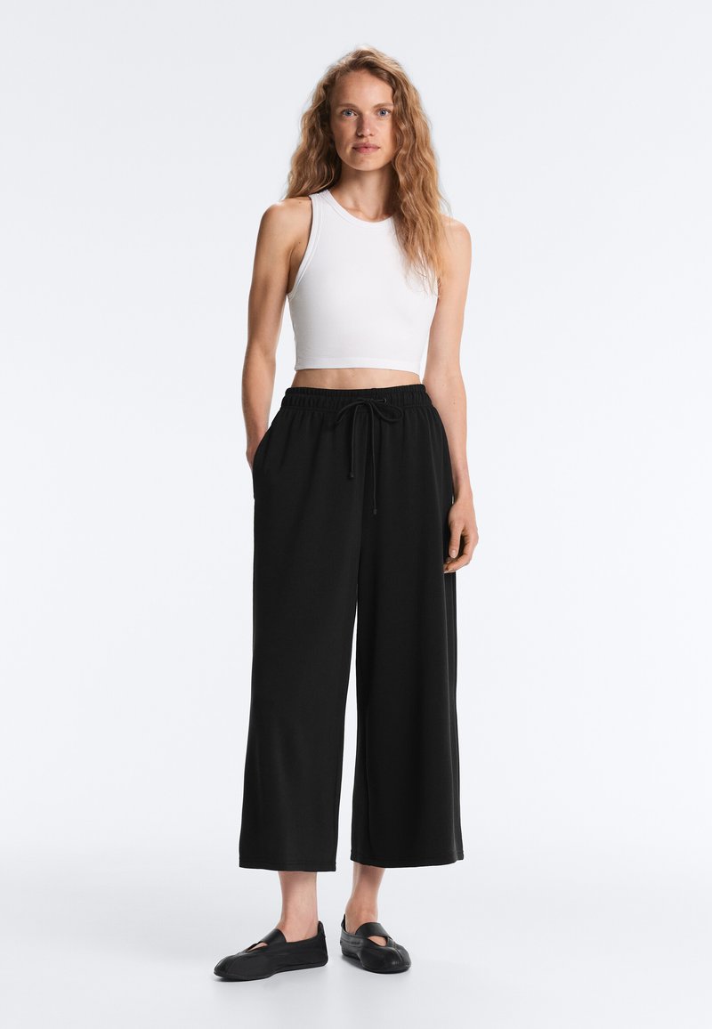 Kobieta z długimi falującymi włosami stoi ubrana w biały, bezrękawnikowy krótki top, luźne czarne spodnie z sznurkiem oraz czarne wsuwane płaskie buty na białym tle.