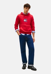 Joven con las manos en los bolsillos, vistiendo una sudadera roja, una camisa de rayas azules, jeans oscuros y zapatos negros, de pie frente a un fondo blanco.