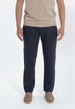 Uomo con camicia beige, pantaloni casual blu scuro e scarpe slip-on marrone chiaro, in piedi davanti a uno sfondo bianco semplice.