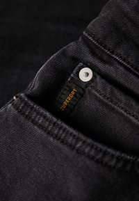 Denim negro con una vista en primer plano de un bolsillo. Presenta un botón plateado y una etiqueta con "Superdry" en texto naranja. Textura suave, diseño minimalista.