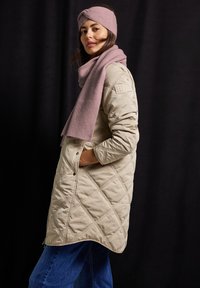 Béžový prošívaný kabát s vysokým límcem, bočními kapsami a zakřiveným lemem. Doplněno o měkký růžový pletený šál a čelenku. Modré jeansy dokončují vzhled.