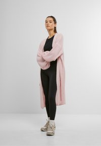 Lichtroze gebreide cardigan met een losse pasvorm, lange mouwen en een open voorkant, gecombineerd met zwarte leggings en lichtgekleurde sneakers.