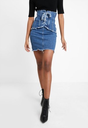 Frau trägt einen High-Waist-Jeanskorsett-Gürtel mit Schnürung über einem Minirock aus Jeans mit ausgefranstem Saum, ein schwarzes Kurzarmshirt und schwarze Schnür-Stiefeletten.