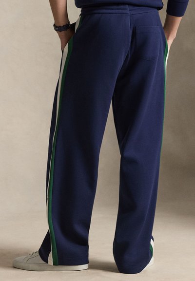 Personne portant un pantalon large bleu marine avec des bandes latérales vertes et blanches et des baskets blanches, mains dans les poches, debout sur un sol beige.