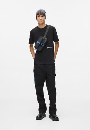 Svart t-skjorte med hvitt logo, svarte cargo-bukser med blå aksent, og en svart crossbody-veske med blå stropper; grå lue og svarte sko.