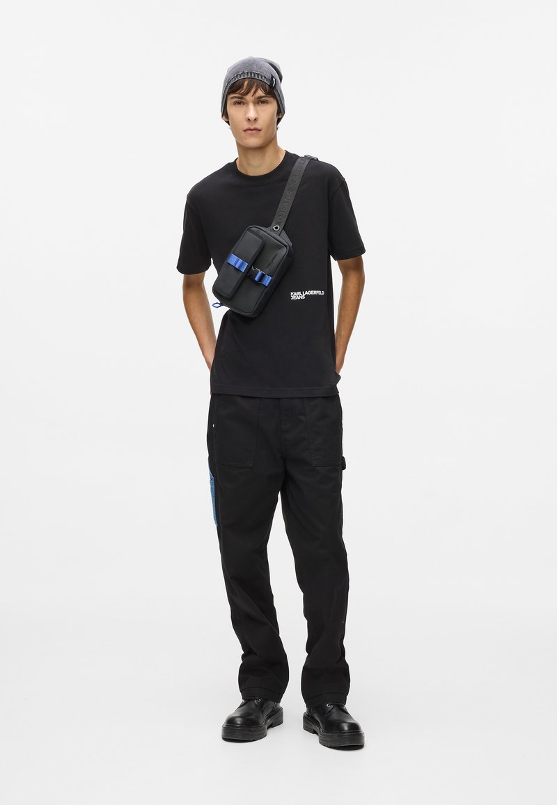 Μαύρη μπλούζα με λευκό λογότυπο, μαύρο παντελόνι cargo με μπλε λεπτομέρεια και μαύρη τσάντα χιαστί με μπλε λουριά; γκρι σκούφος και μαύρα παπούτσια.