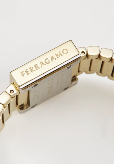 Montre en métal doré avec un boîtier rectangulaire et un bracelet à maillons. Présente le logo gravé "FERRAGAMO", une finition lisse et une surface polie.