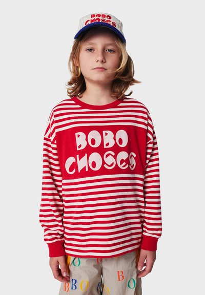 Rot-weiß gestreifte Langarmshirt mit dem Aufdruck "BOBO CHOSES". Das Modell trägt eine passende Mütze mit blauem Schirm und bunten Buchstaben.