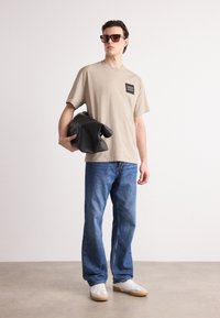 Tricou de bumbac bej cu un petic pătrat negru, asortat cu blugi din denim albastru și adidași albi. Modelul ține o geantă neagră.