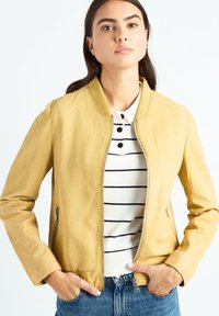 Veste aviateur en cuir jaune avec un col côtelé, une fermeture éclair devant et des poches latérales, associée à une chemise blanche rayée et un jean en denim.