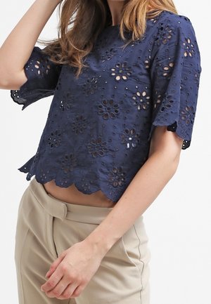 Nainen, jolla on tummansininen lyhythihainen pusero, jossa on kukkakuvioisia neulontakoreja ja lomittain aaltoilevat reunat, yhdistettynä beigeihin korkeavyötäröisiin housuihin.