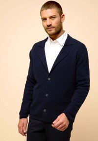  Marinblå cardigan i texturerat tyg, med sjalkrage, knappfram och långa ärmar, bärs över en vit skjorta.