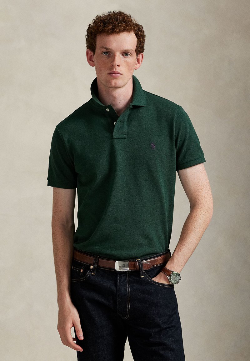 Jeune homme aux cheveux roux bouclés portant un polo vert foncé, un jean foncé, une ceinture marron et une montre-bracelet, debout devant un fond uni.
