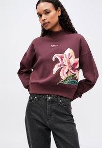 Mujer vistiendo un sudadera burdeos con un gran estampado de flores en rosa y blanco, y jeans de tiro alto negros, contra un fondo sencillo.