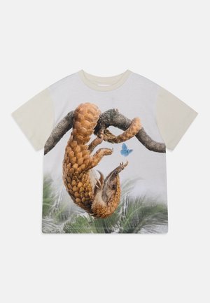 Chemise blanche à manches courtes avec des manches beige clair montrant un pangolin suspendu à une branche regardant un papillon bleu.