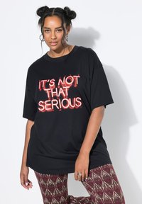 Zwarte oversized t-shirt met rode neon tekst "HET IS NIET ZO SERIEUS." Glad stof, comfortabele pasvorm. Gecombineerd met gemusterde leggings.