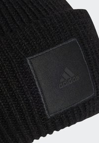 Svart stickad mössa med ribbad textur. Har ett rektangulärt svart tygmärke med en prägling av logotypen med tre ränder.