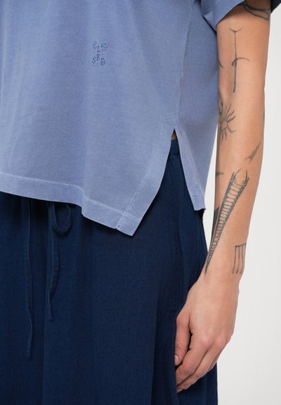 Φωτεινό γαλάζιο βαμβακερό μπλουζάκι με κοντά μανίκια και πλαϊνές σχισμές, συνδυασμένο με μια φούστα σε σκούρο μπλε χρώμα με κορδόνι στη μέση. Διαθέτει κεντημένο λογότυπο.