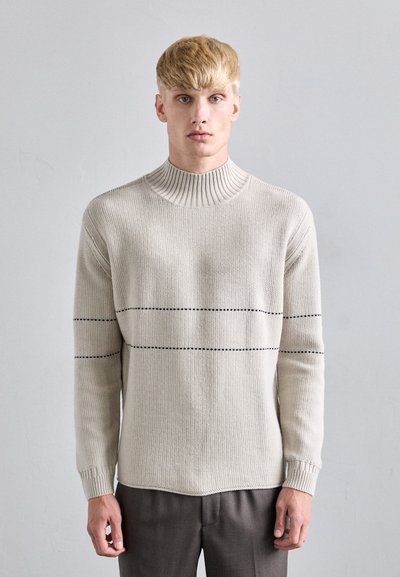 Suéter de punto beige claro con un alto cuello acanalado, mangas largas y rayas negras horizontales en el pecho. Textura suave.