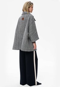 Grijze houndstooth-gepatenteerde jas met een brede silhouet, grote mouwen en een leren label op de achterkant, gecombineerd met marineblauwe wijde broeken.