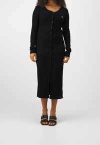 Musta napillinen mekko, jossa on pitkät hihat, V-pääntie ja pehmeä rakenne. Sisältää etupuolella halkion ja koristeellisen logon vasemmassa rinnassa.