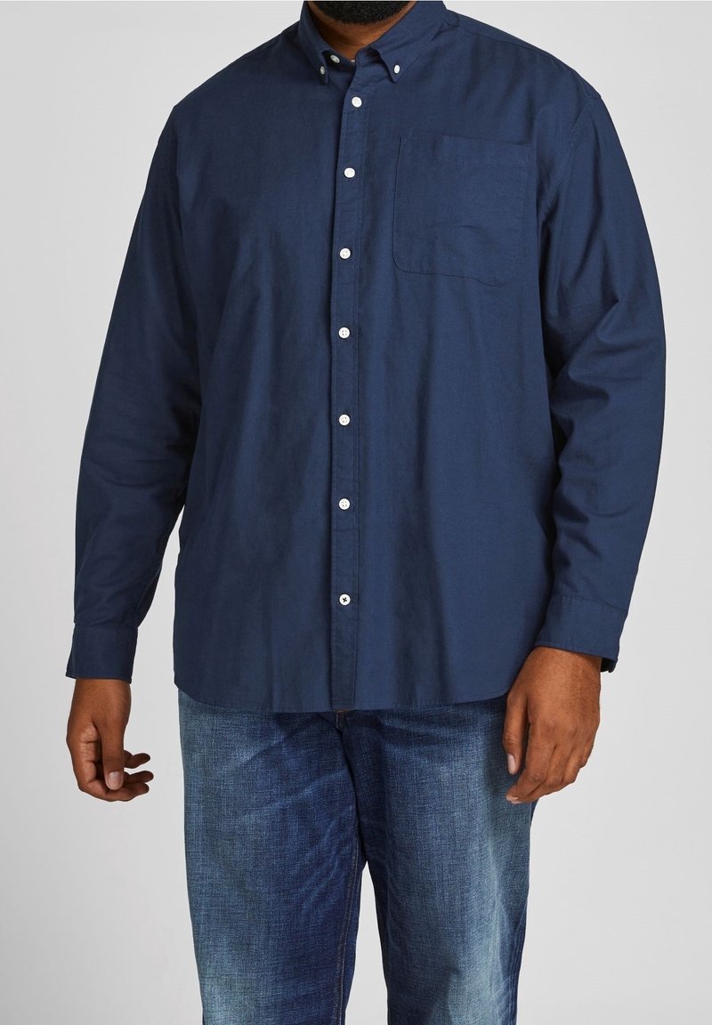 Homme portant une chemise bleu foncé à manches longues et un jean bleu délavé moyen, debout devant un fond uni.