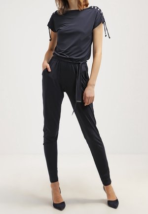 Femme portant une combinaison noire ajustée avec des détails à lacets sur les épaules et une ceinture nouée à la taille, assortie à des talons pointus noirs.