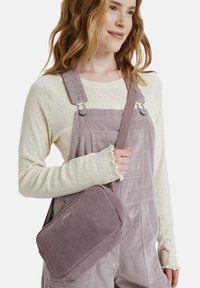 Violetti corduroy-olkalaukku, jossa on suorakaiteen muotoinen, tekstuuripinta ja säädettävä hihna, jota kantaa päällä vaalean kermanvärinen teksturoitu pitkähihainen paita.