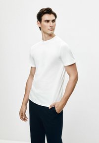 Biała koszulka z krótkim rękawem, wykonana z bawełny, z okrągłym dekoltem i dopasowanym krojem, w połączeniu z ciemnymi spodniami, na neutralnym tle.