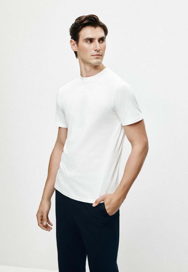 Biała koszulka z krótkim rękawem, wykonana z bawełny, z okrągłym dekoltem i dopasowanym krojem, w połączeniu z ciemnymi spodniami, na neutralnym tle.