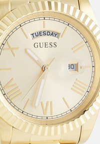 Montre Guess dorée avec un cadran texturé, marqueurs d'heure blancs, fonction date et affichage du jour indiquant "MARDI" sur un cadran doré clair.