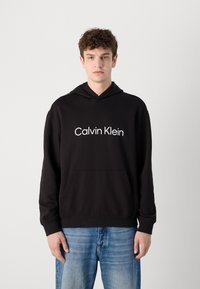 Juodas hoodie'as su priekiu kišenėmis, kuriame yra balti "Calvin Klein" užrašai. Sukurtas su standartiniu kirpimu ir taip pat turi raukšles ant rankovių. Dėvimas su mėlynomis džinsinėmis kelnėmis.