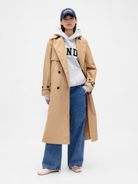 Beež trench coat kahekordse nööpimisega halli kapuutsiga jakka üle, koos laia säärega siniste teksade ja heledate lühikeste kingadega; tumesinine müts, millel on tekst.