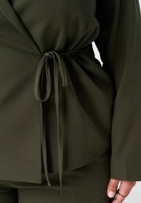 Olijfgroene top van zachte stof met een gerimpelde tailleband die is vastgebonden in een strik. Lange mouwen met een losse pasvorm en een gladde textuur.