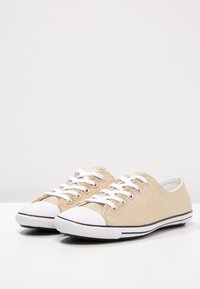 Zapatillas de lona beige con punteras y cordones de goma blanca, que cuentan con una franja negra a lo largo de la suela y una parte superior texturizada.