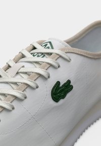 Λευκό αθλητικό παπούτσι με μπεζ περιθώριο, λευκά κορδόνια και πράσινο λογότυπο κροκόδειλου στο πλάι, σε απλό φόντο.