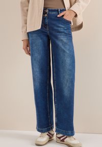 Jean bleu à jambe large en denim, avec deux fermetures à bouton, de grandes poches et un ourlet effiloché. Associé à des baskets blanches et bordeaux.